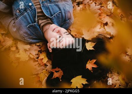 Asiatische Frau liegt in einem Haufen von herbstlich orangefarbenen und gelben Blättern Stockfoto