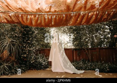 Schönes Hochzeitskleid mit Falten und Schwanz hängen auf weißen Kleiderbügel im Freien. Ballkleid im Garten. Stockfoto