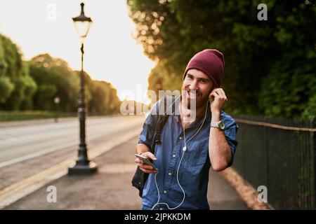 Die Musik macht mich bewegt. Ein beschnittenes Porträt eines hübschen jungen Mannes, der Musik auf seinem Handy hört, während er durch die Stadt läuft. Stockfoto