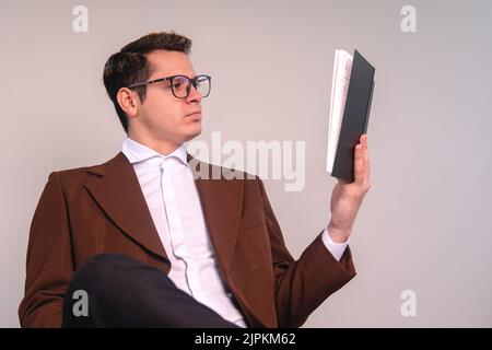 Ein Universitätsprofessor auf einem isolierten weißen Hintergrund, der aufmerksam ein Buch liest. Ein eleganter Mann, der studiert. Junger Mann in braunem Anzug und Brille. Hochwertige Fotos Stockfoto