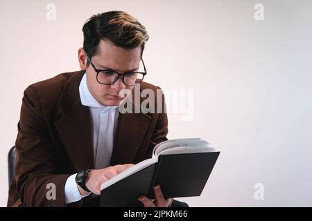 Intelligent gekleideter studious Mann. Mann in brauner Jacke, weißem Hemd, Brille und auf weißem Hintergrund. Geschäftsmann liest ein schwarzes Buch. Rechtsanwalt, der mit seinen Notizen arbeitet. Hochwertige Fotos Stockfoto