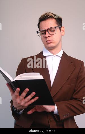 Verwirrt eleganter Mann aufmerksam ein Buch zu lesen. Ein Mann in Anzug, Hemd und Brille beim Studieren. Ein studious Mann, der sein schwarzes Buch liest. Geschäftsmann analysiert aufmerksam sein Buch. Hochwertige Fotos Stockfoto