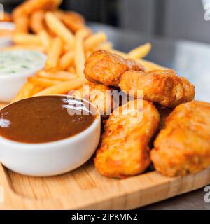 Eine Nahaufnahme von köstlichen Chicken Nuggets und Pommes frites, serviert mit Saucen auf einem Holzbrett Stockfoto