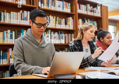 Nichts kann ihm die Konzentration nehmen. Ein fokussierter junger Mann arbeitet an seinem Laptop und studiert, während er in einer Bibliothek sitzt. Stockfoto