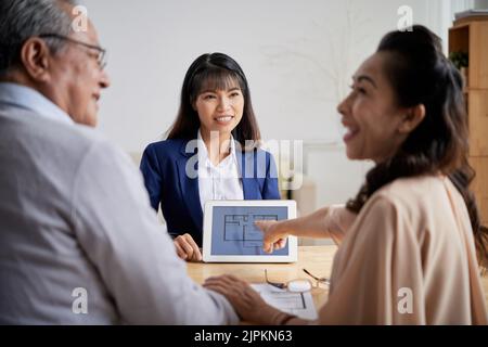 Ziemlich junge Immobilienmaklerin, die ihren älteren Kunden einen Apartment-Plan zeigt Stockfoto