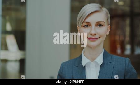 Nahaufnahme des Porträts einer erfolgreichen blonden Geschäftsfrau, die lächelt und in die Kamera schaut, in einem modernen Büro im Innenbereich Stockfoto