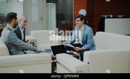 Fröhlicher Geschäftsmann, der Finanzberichte mit Geschäftspartnern diskutiert und auf dem Sofa in der modernen Bürohalle sitzt Stockfoto
