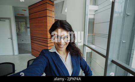 POV der jungen Geschäftsfrau im Anzug, die ein Selfie-Foto mit dem Smartphone hält und Spaß im modernen Büro im Innenbereich hat Stockfoto