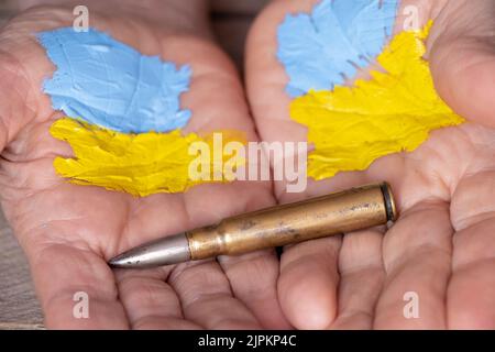 Die Hände einer alten Frau mit einer bemalten Flagge der Ukraine halten eine militärische Kugel auf einem hölzernen Hintergrund, Krieg in der Ukraine, Frau Militär Stockfoto