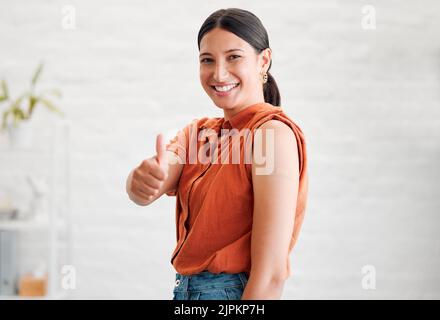 Positive junge Frau, die mit einer Covid-Impfstoffinjektion behandelt wurde, zeigt mit Daumen nach oben und zeigt ihren Arm, um eine Immunisierung zu genehmigen. Glückliches, gesundes Weibchen Stockfoto