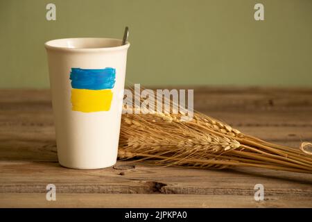 Die gelb-blaue Flagge der Ukraine ist auf einer weißen Tasse mit Kaffee auf dem Tisch in der Nähe des Weizenfeldes in der Ukraine gemalt, den Krieg zu stoppen Stockfoto