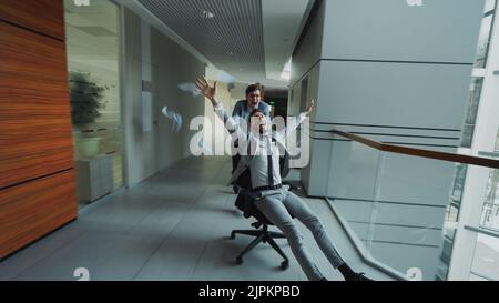 Zwei verrückte Geschäftsleute reiten auf einem Bürostuhl und werfen Zeitungen, während sie sich in der Lobby des modernen Business-Centers im Innenbereich Vergnügen Stockfoto