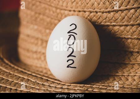 Weißes Hühnerei auf schwarzem Hintergrund mit geschriebenem Text 2022 liegt auf einem gelben Strohhut, ostern und Kultur im neuen Jahr, Essen Stockfoto