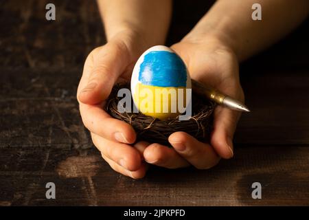 Ein weißes Hühnereier mit einer bemalten ukrainischen Flagge in einem Strohkorb und einer Kugel in den Händen einer Frau auf einem Tisch zu Hause, die ihr Haus beschützte 20 Stockfoto