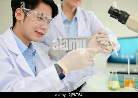 Ein intelligenter, tausendjähriger asiatischer Wissenschaftler passt in seinem Wissenschaftslabor zusammen mit seiner leitenden weiblichen Vorgesetzten eine Virusprobe in einer Petrischale an. Pharmakologie Stockfoto