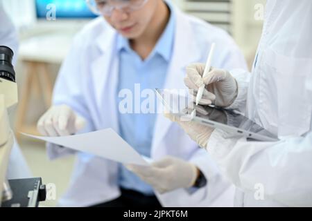 Eine Wissenschaftlerin, die mit ihrer Kollegin im Labor arbeitet und die Ergebnisse des Experiments auf dem Tablet-Touchpad aufzeichnet Stockfoto