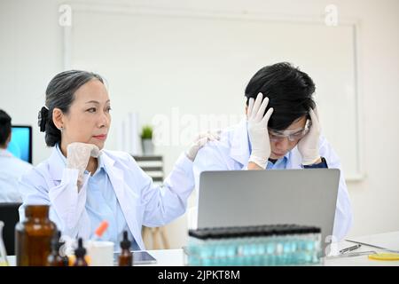 Gestresster junger asiatischer Wissenschaftler verärgert und unzufrieden mit seinem medizinischen Experiment-Projekt, während er im Labor mit einer älteren Professorin arbeitete. Stockfoto
