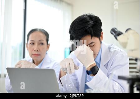 Gestresster junger asiatischer Wissenschaftler, der mit seinem Kollegen im Labor arbeitete, verärgert über sein Chemieexperiment-Projekt. Stockfoto