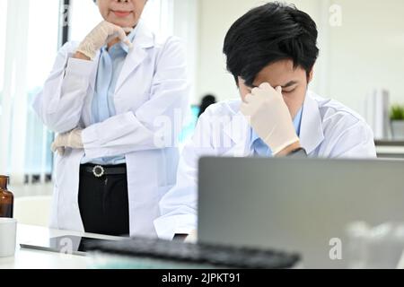 Unglücklicher und gestresster junger asiatischer Wissenschaftler sitzt an seinem Schreibtisch im Labor, nachdem er eine Beschwerde von seiner weiblichen Vorgesetzten erhalten hat. Stockfoto