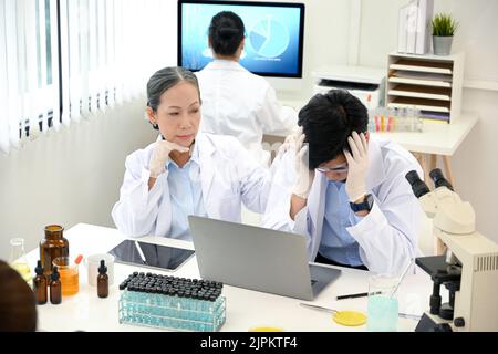 Gestresster und ernsthafter, tausendjähriger asiatischer Wissenschaftler verärgert über sein medizinisches Experimentierprojekt, während er im Labor mit einer höheren Professorin arbeitete. Stockfoto