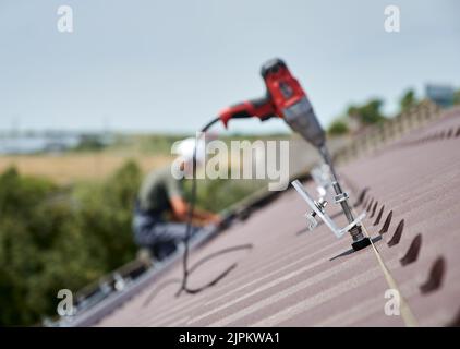 Nahaufnahme der Klemme. Arbeiter Vorbereitung auf die Installation von Photovoltaik-Solarpanel-System auf dem Dach des Hauses. Mann und elektrischer Schraubendreher auf unscharfem Hintergrund. Stockfoto