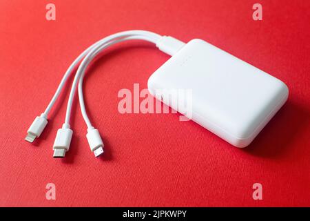 Weiße Powerbank auf rotem Hintergrund. Powerbank mit verschiedenen Steckverbindern. Externer Akku für verschiedene Geräte. Stockfoto