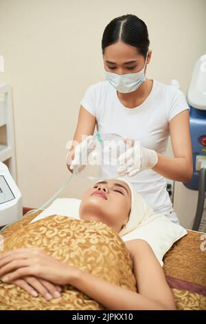 Hochprofessionelle junge Kosmetikerin mit Gummihandschuhen und medizinischer Gesichtsmaske, die am Behandlungstisch steht, während sie sich auf die Arbeit mit speziellen Geräten konzentrierte Stockfoto