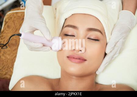 Nahaufnahme der Hände der Kosmetikerin, die eine Ultraschallbehandlung durchführen, während ein hübscher asiatischer Klient mit geschlossenen Augen auf dem Behandlungstisch liegt Stockfoto