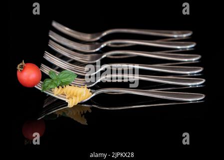 Eine große Metallgabel mit Nudeln, Basilikum und einer Tomate darauf. Im Hintergrund befinden sich leere Gabeln in einer Reihe vor dunklem Hintergrund. Stockfoto