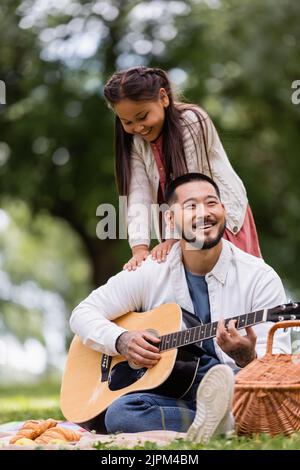 Asiatisches Kind, das beim Picknick im Park neben dem Vater steht und akustische Gitarre spielt Stockfoto