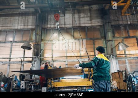 Arbeiter heben Metallblech mit Krankettenzug mit Fernbedienung und Haken in der industriellen Fabrik Stockfoto