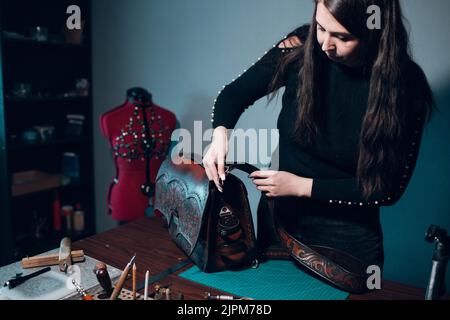 Tanner Frau macht Nähtasche mit Musterlederwaren in der Werkstatt. Arbeitsprozess des Lederhandwerks. Stockfoto