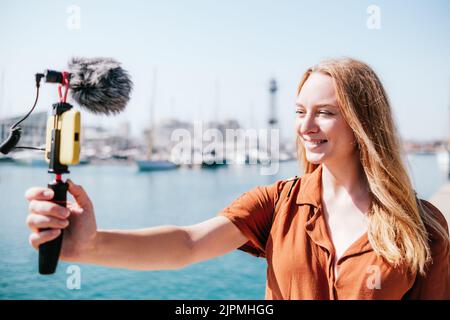 Weibliche Reise-Vlogger Streaming in der Nähe von Meer in einem touristischen Ziel. Sie schaut lächelnd auf ihr Smartphone Stockfoto