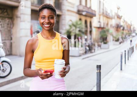 Horizontales Porträt einer positiven afroamerikanischen Frau, die auf der Straße die Kamera anschaut. Sie lächelt und hält einen wiederverwendbaren Kaffeebecher und ein rotes Telefon Stockfoto