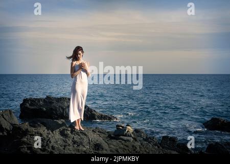 Die ganze Länge der friedlichen jungen barfüßigen Dame mit langen dunklen Haaren in maxi weißem Kleid steht auf rauem felsigen Felsbrocken und blickt hinunter in die Nähe des welligen Meeres A Stockfoto