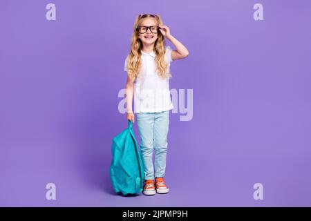 Ganzkörper-Foto von kleinen Mädchen Pferdeschwänze berühren Brillen Rucksack bereit Schule gekleidet stilvollen weißen Blick isoliert auf violetten Hintergrund Stockfoto