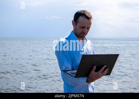 Ein Mann mit einem Laptop im Meer in einem weißen Bürohemd. Arbeiten im Urlaub. Stockfoto