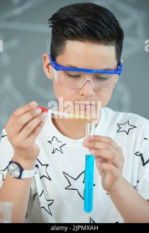 Hübscher asiatischer Teenager, der eine Schutzbrille mit Reagenzglas und Pipette trägt, während er im Klassenzimmer der Chemie experimentiert, Porträtaufnahme Stockfoto