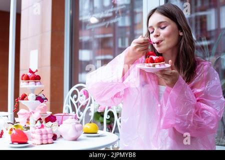 Hübsches Teenager-Mädchen in rosa Regenmantel joying, Verkostung süße Beere Dessert Kuchen vom Tisch im Café. Geburtstagsereignis Stockfoto