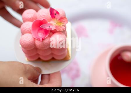 Nahaufnahme pinkfarbener neo Cube Ball Velours Mousse Kuchen Dessert mit Blütenblättern und Schmetterling auf dem Teller in den Händen in der Nähe einer Tasse Tee Stockfoto