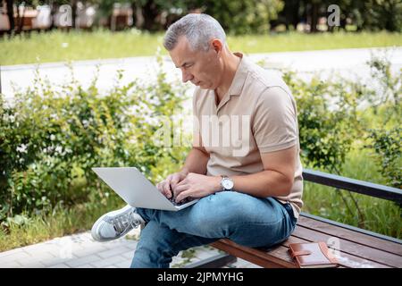 Porträt eines konzentrierten älteren Mannes, der im Sommer auf einer Bank mit gekreuzten Beinen im Park sitzt und am Laptop tippt. Stockfoto