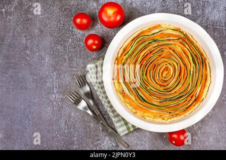 Hausgemachte, spiralförmige Gemüsetorte aus Karotten, Zucchini und Auberginen mit Bechamelsauce auf grauem Betongrund. Rustikaler Stil, Draufsicht, flaches Lay Stockfoto