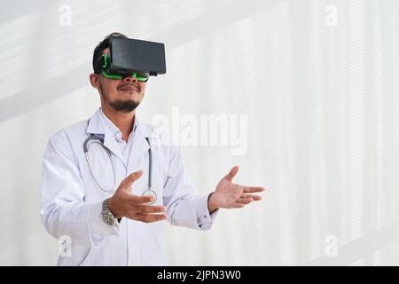 Bärtiger Arzt mit weißem Mantel und VR-Headset bei der Arbeit im modernen Büro, weißer Hintergrund Stockfoto