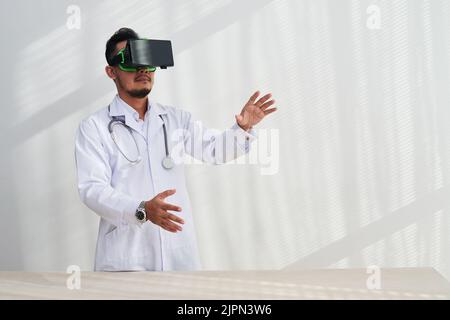 Hochprofessioneller Arzt mit weißem Mantel und VR-Headset, der Fernkonsultation durchführt, während er im modernen Büro steht Stockfoto