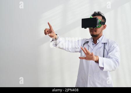 Hochprofessioneller Arzt mit Virtual-Reality-Headset, der Krankenakten überprüft, während er in einem modernen Büro mit weißem Hintergrund arbeitet Stockfoto