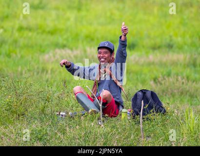 Ein fröhlicher indonesischer Mann mittleren Alters, der mit dem Daumen nach oben auf Gras sitzt. Sulawesi, Indonesien. Stockfoto