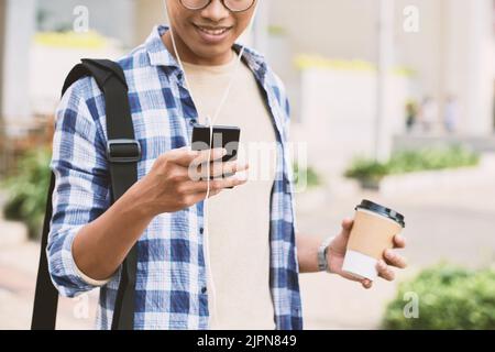 Nahaufnahme eines nicht erkennbaren jungen Mannes, der Kopfhörer trägt und mit einem Freund auf dem Smartphone SMS schreibt, während er im Freien steht und duftenden Kaffee und verschwommenen Hintergrund genießt Stockfoto