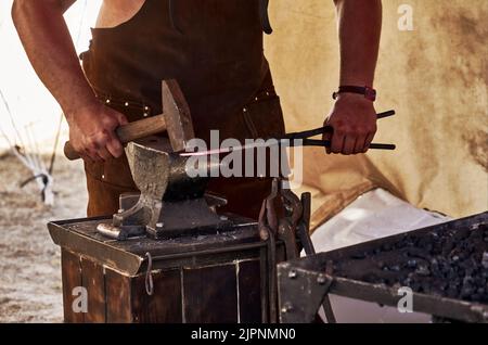 Der Schmied in einem Ledermantel arbeitet mit einem Hammer auf einem Amboss ein rotglühend heißes Eisen Stockfoto