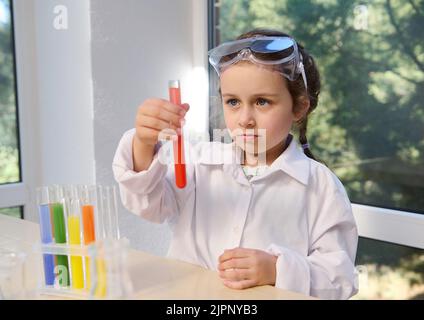 Mädchen im Kindergarten mit Schutzbrille und Laborkittel, die eine chemische Reaktion im Reagenzglas beobachtet, die sie in der Hand hält. Stockfoto