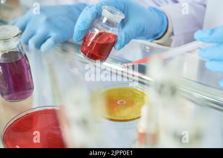 Nahaufnahme eines nicht erkennbaren Wissenschaftlers, der mit einer Pipette Gummihandschuhe trägt, während er zwei Flüssigkeiten in einer Petrischale mischt, wobei sich Laborglas im Vordergrund befindet Stockfoto